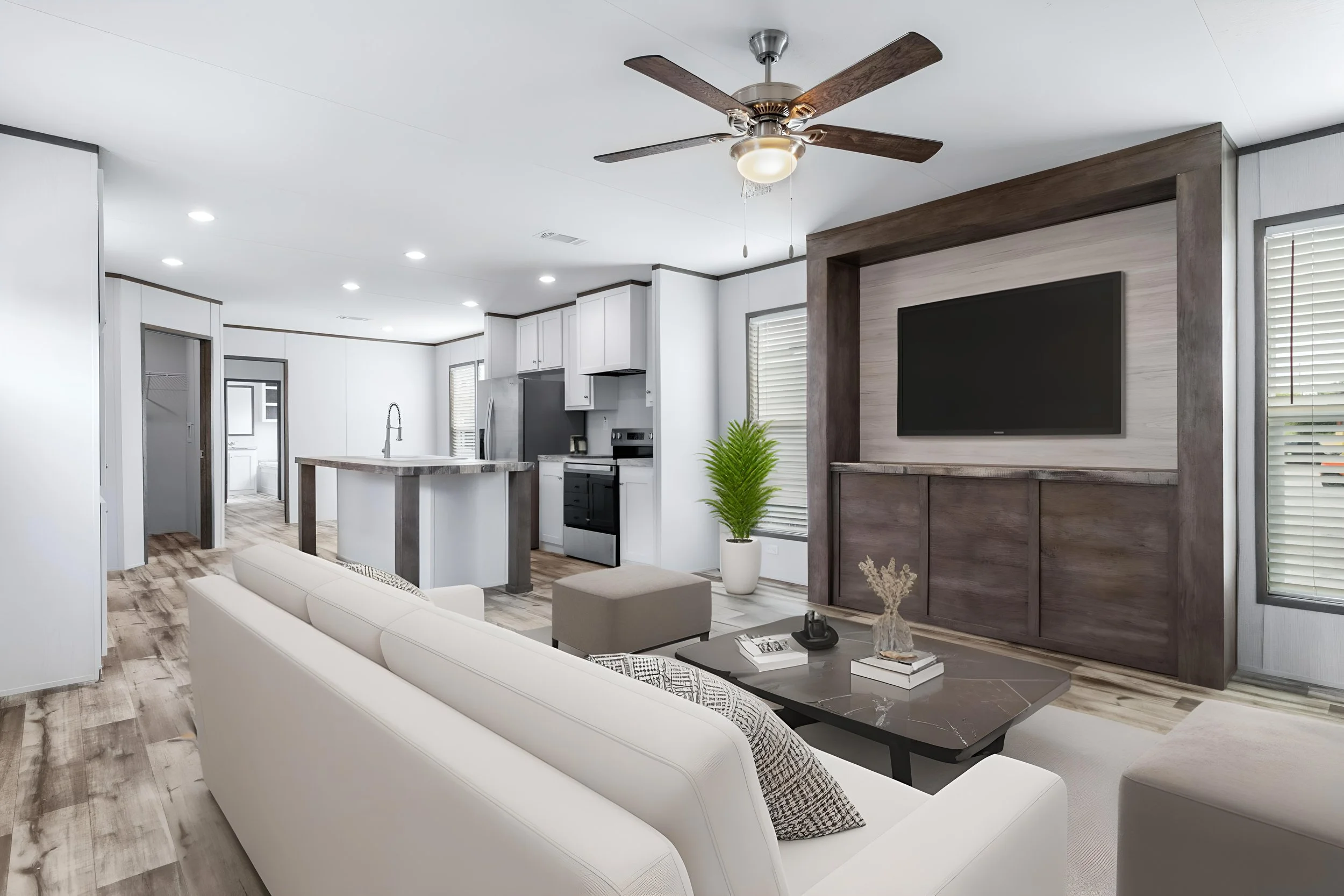 Modern open-concept living room and kitchen with white walls, wood flooring, a ceiling fan, a wall-mounted flat-screen TV, and a kitchen area with white cabinets and stainless steel appliances.