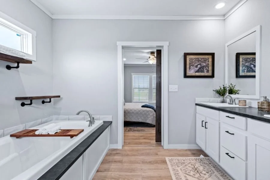 Bathroom with white vanity, large mirror, potted plants, framed pictures, bathtub with wooden tray and towels, wood flooring, view into bedroom with bed and windows.
