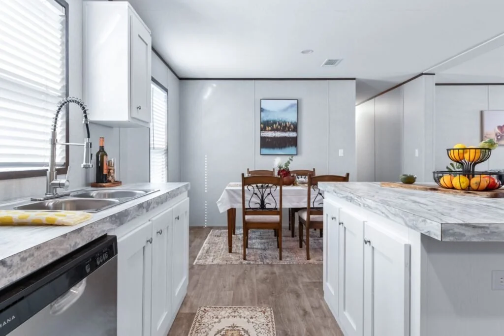 Modern kitchen and dining area with white cabinets, marble countertops, a stainless steel sink, and wooden dining table with four chairs. There is a framed landscape on the wall and a fruit basket on the counter.