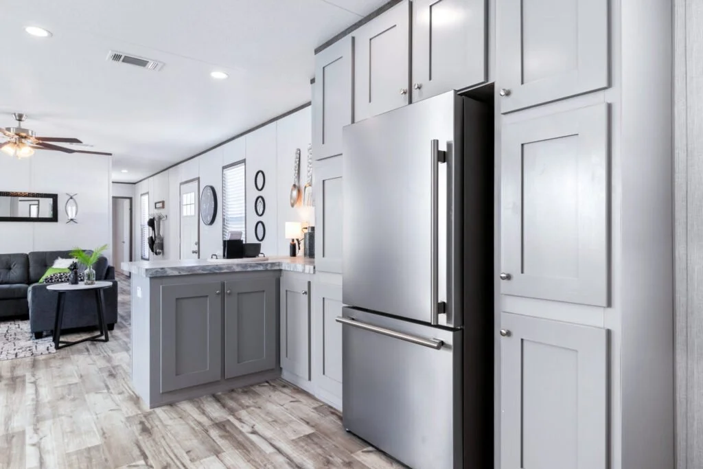 Modern kitchen with gray cabinets and stainless steel refrigerator, open to a living room with a black sofa and decorative wall art.