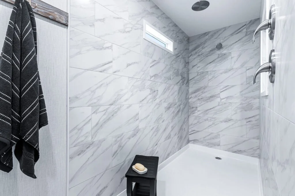 Bathroom shower with marble tiles, a small window, a black shower stool with soap, and two shower controls.