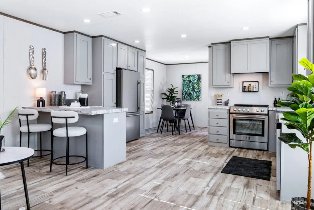 A modern kitchen with gray cabinets, stainless steel appliances, a small bar area with two chairs, a dark dining table with four chairs, and decorative plants.
