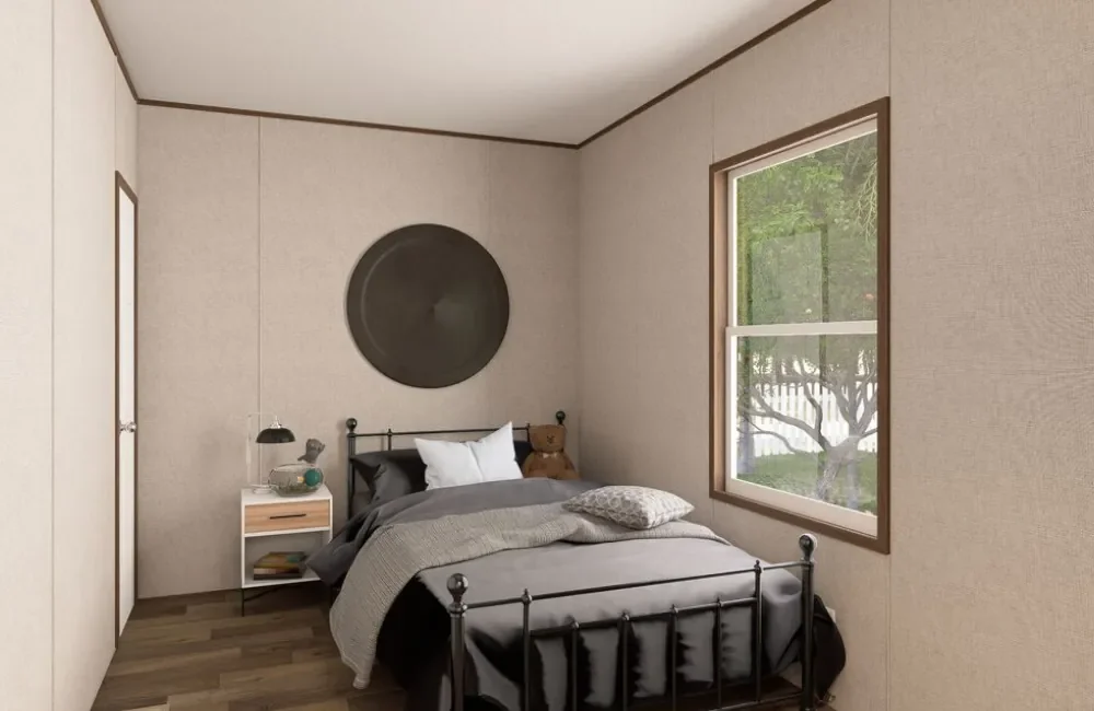 A small bedroom with beige walls, a black metal bed, a nightstand with a lamp, teddy bear, and books, a window showing trees outside, and a round wall decoration above the bed.