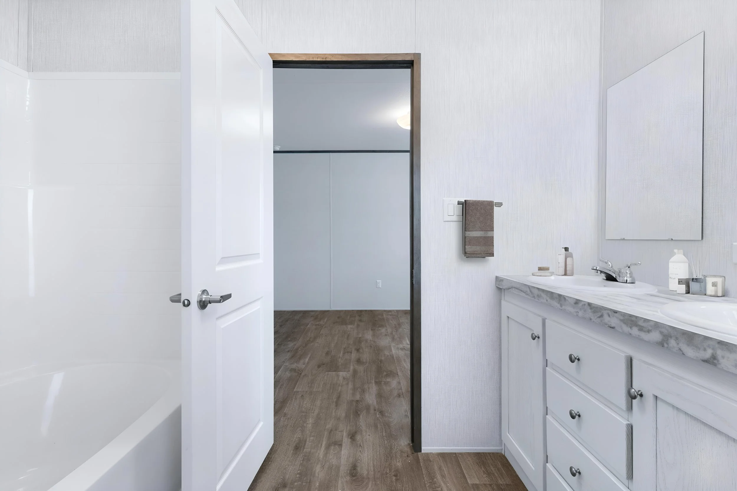 A clean modern bathroom with a white cabinet and marble countertop, a rectangular mirror, and various toiletries on the counter. The image is taken from an open door with a white frame, showing a wood floor and a light-colored wall.