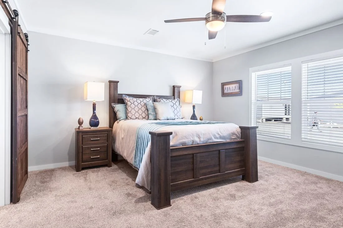 Bedroom with a wooden bed frame, beige bedding, and multiple pillows, two nightstands with lamps, carpeted floor, two windows with blinds, a framed picture on the wall, ceiling fan, and light-colored walls.