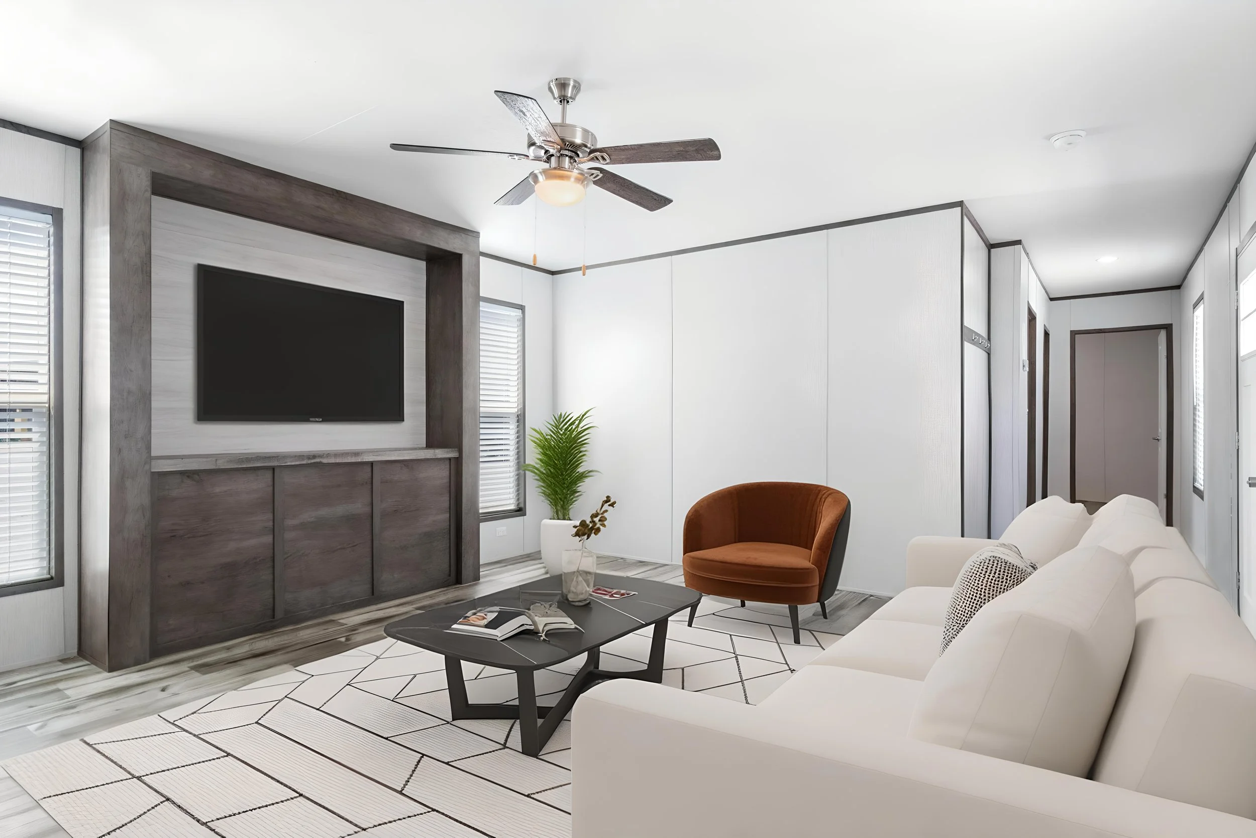 Modern living room with white walls, a flat-screen TV, a ceiling fan, a white sofa, a brown armchair, a black coffee table, and a geometric rug. There are windows with blinds and some indoor plants.
