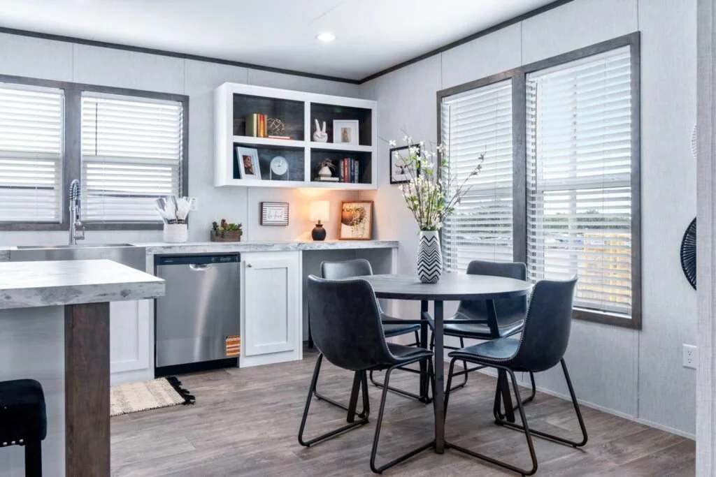 A modern kitchen and dining area with gray and white color scheme, featuring a round dining table with four black chairs, large windows with blinds, open shelving with decor items, and a small lamp on the countertop.
