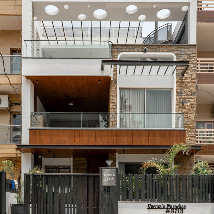 Modern multi-story building with balconies, glass railings, wooden accents, and stone facade, featuring a sign that reads "Verma's Paradise" at the entrance.