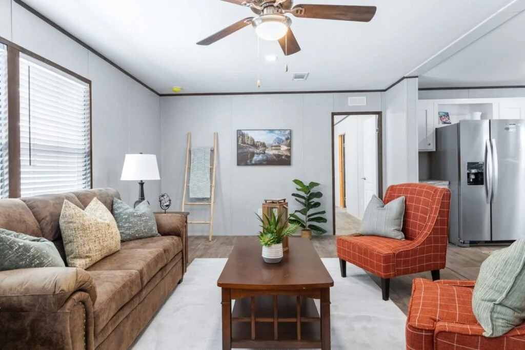 Living room with brown sofa and patterned pillows, orange armchairs with pillows, wooden coffee table with plant, white walls, ceiling fan, views into kitchen with stainless steel refrigerator, window with blinds, and framed landscape painting.