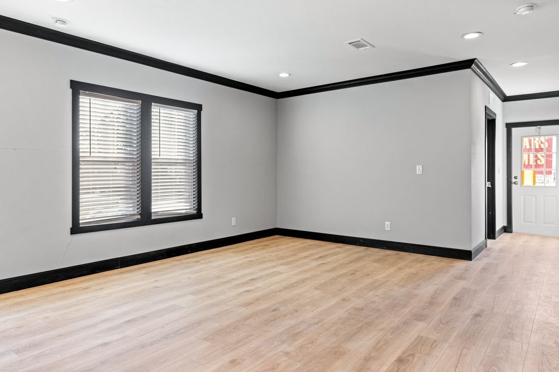 Empty living room with gray walls, black trim, wooden floor, and windows with blinds.