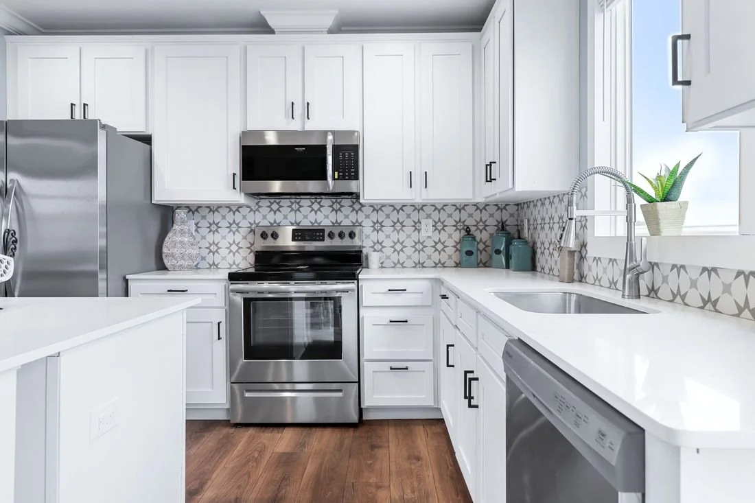 A modern kitchen with white cabinets, a stainless steel refrigerator, microwave, oven, and dishwasher. The backsplash features a gray geometric pattern, and the countertops are white. There is a window with a potted plant and some teal canisters on t