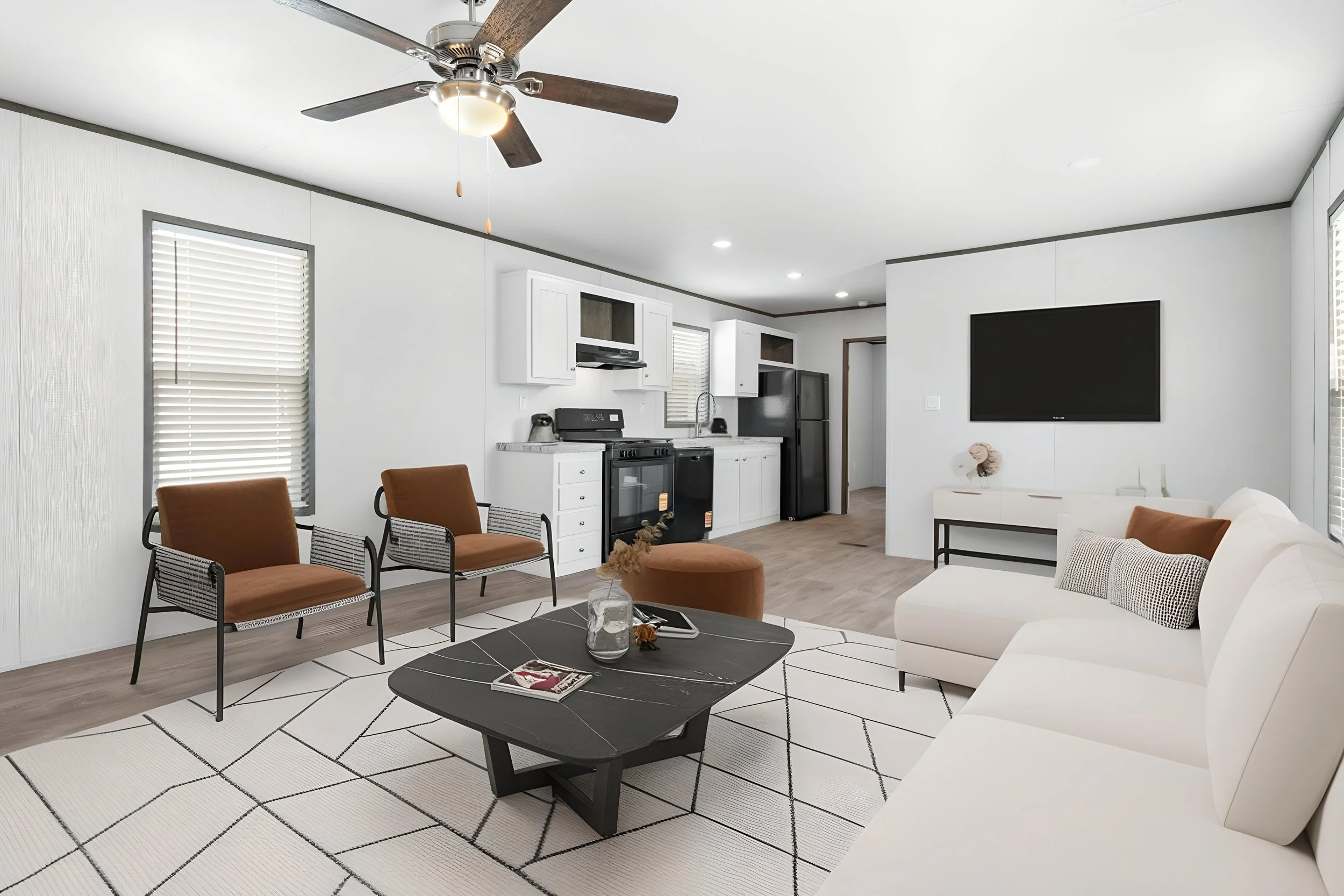 Modern living room with a white sofa, brown chairs, black coffee table, wall-mounted TV, kitchen area with white cabinets, black appliances, and a ceiling fan, bright natural light from windows with blinds, and a light-colored wood floor.