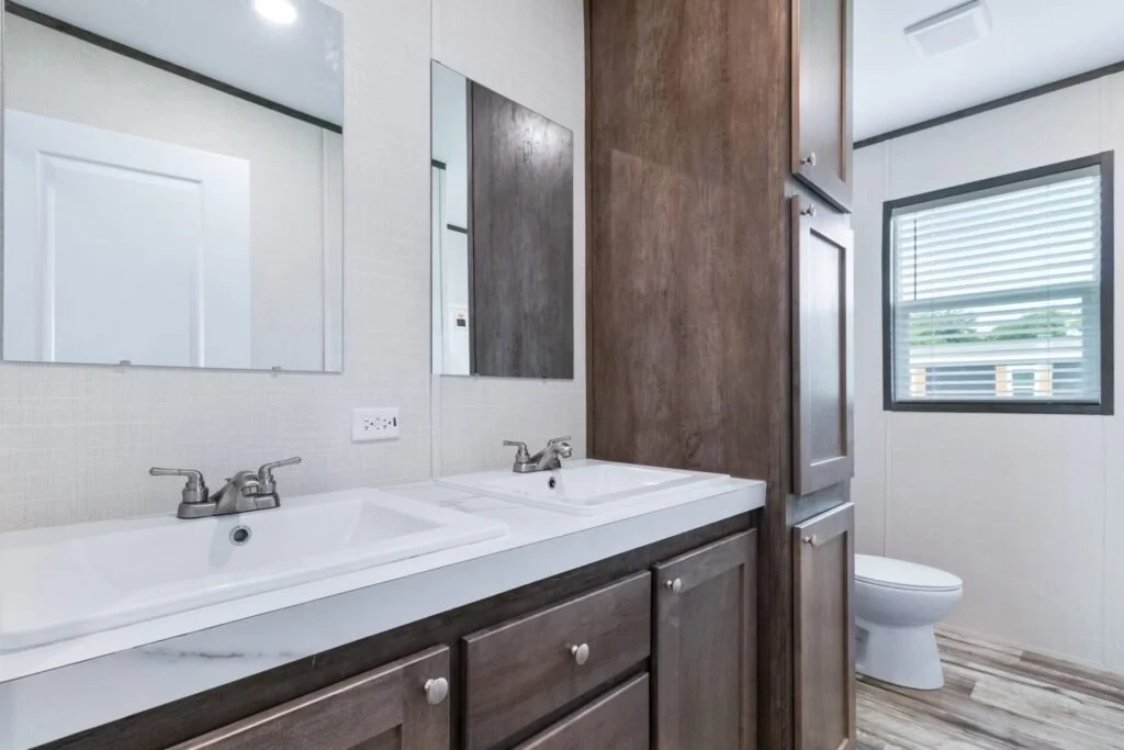 Bathroom with double sinks, mirrors, wooden cabinets, a toilet, and a window with blinds.