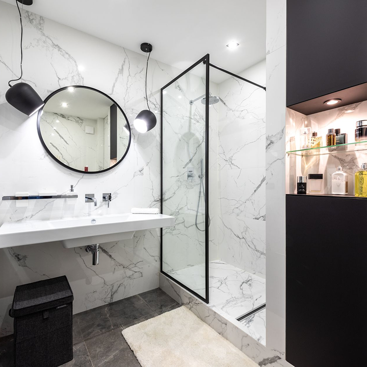 Modern bathroom with marble walls, a round mirror, a white sink, black hanging lights, a walk-in glass shower with marble tiles, and a black shelving unit with toiletries.