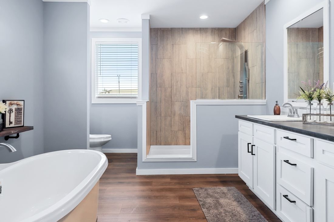 Modern bathroom with a white bathtub, a vanity with a black countertop, a large mirror, a glass-enclosed walk-in shower with wood-look tiles, a toilet, a window with blinds, and wooden flooring.