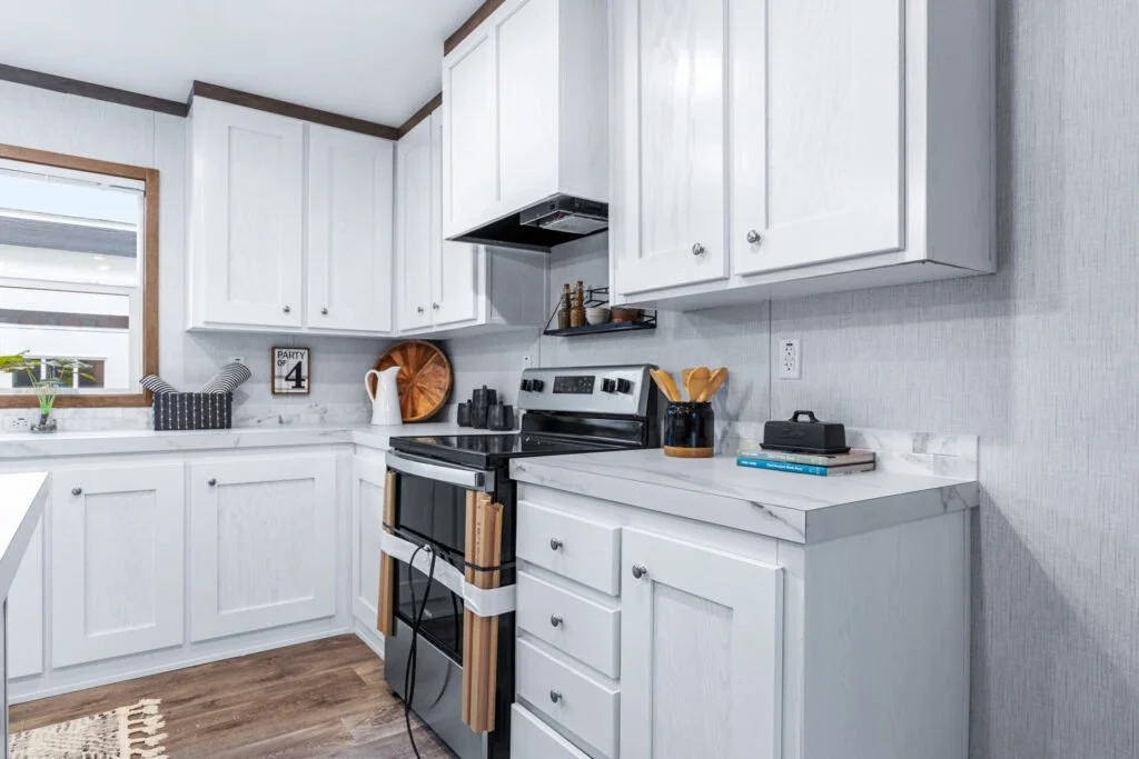 White kitchen with cabinets, a window with blinds, a stove, and countertop accessories including a white pitcher, a wooden tray, black spice jars, a utensil holder with wooden spoons, and books. The flooring is wood, and there is a small sign on the 