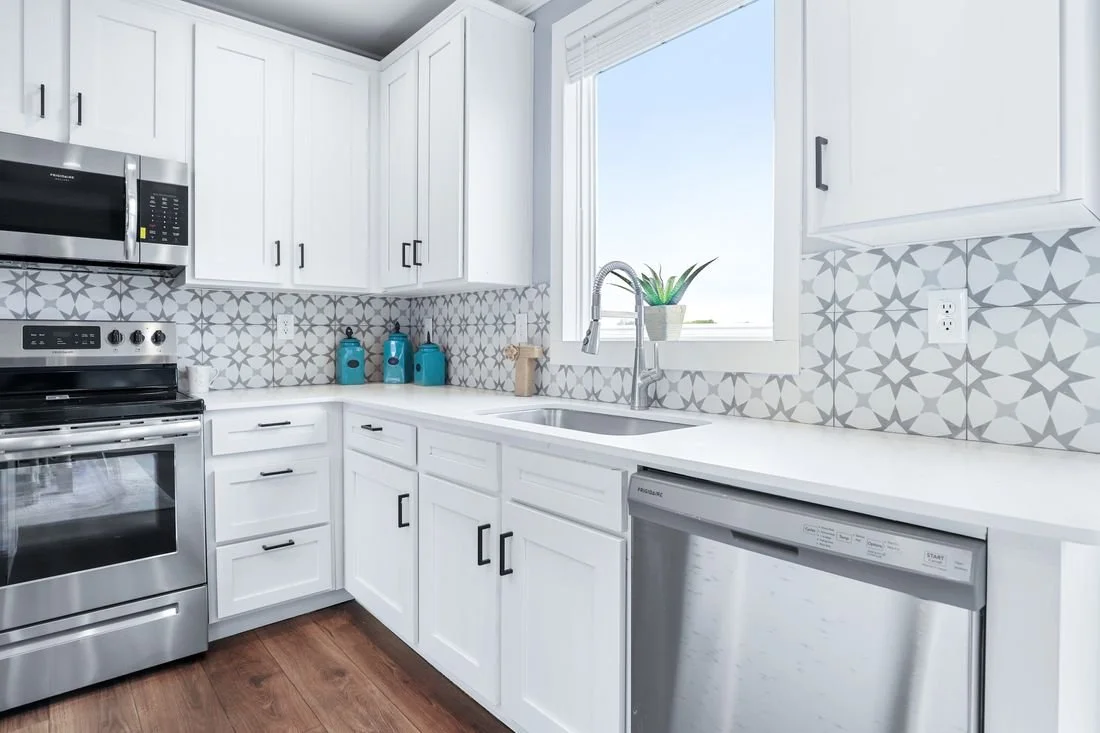Modern kitchen with white cabinets, gray patterned backsplash, stainless steel appliances, a window with blinds, and a small potted plant by the sink.