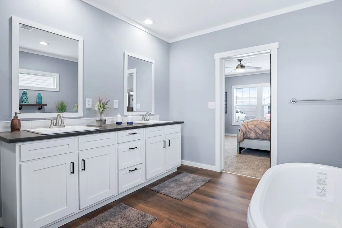 Bathroom with dual sinks, large mirrors, white cabinets, grey walls, hardwood flooring, and a doorway leading to a bedroom with a bed and window.