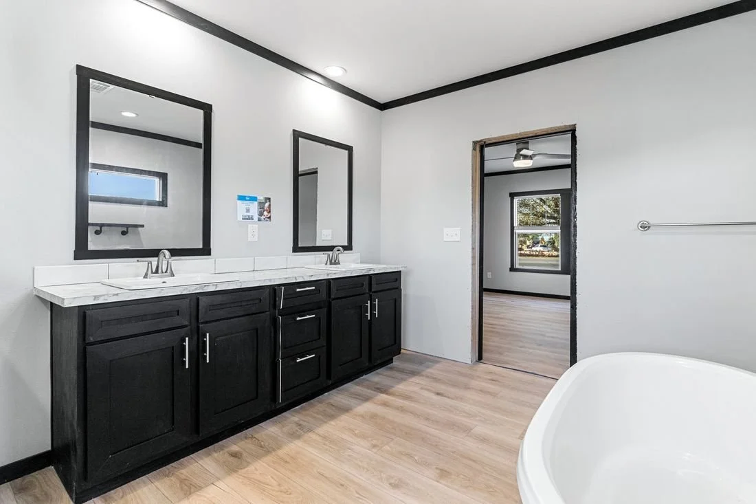 Bathroom with double sinks, black cabinet vanity, white countertop, two mirrors, light-colored wood flooring, doorway leading to another room, partial view of a bathtub, window with gray trim, ceiling fan in the adjacent room