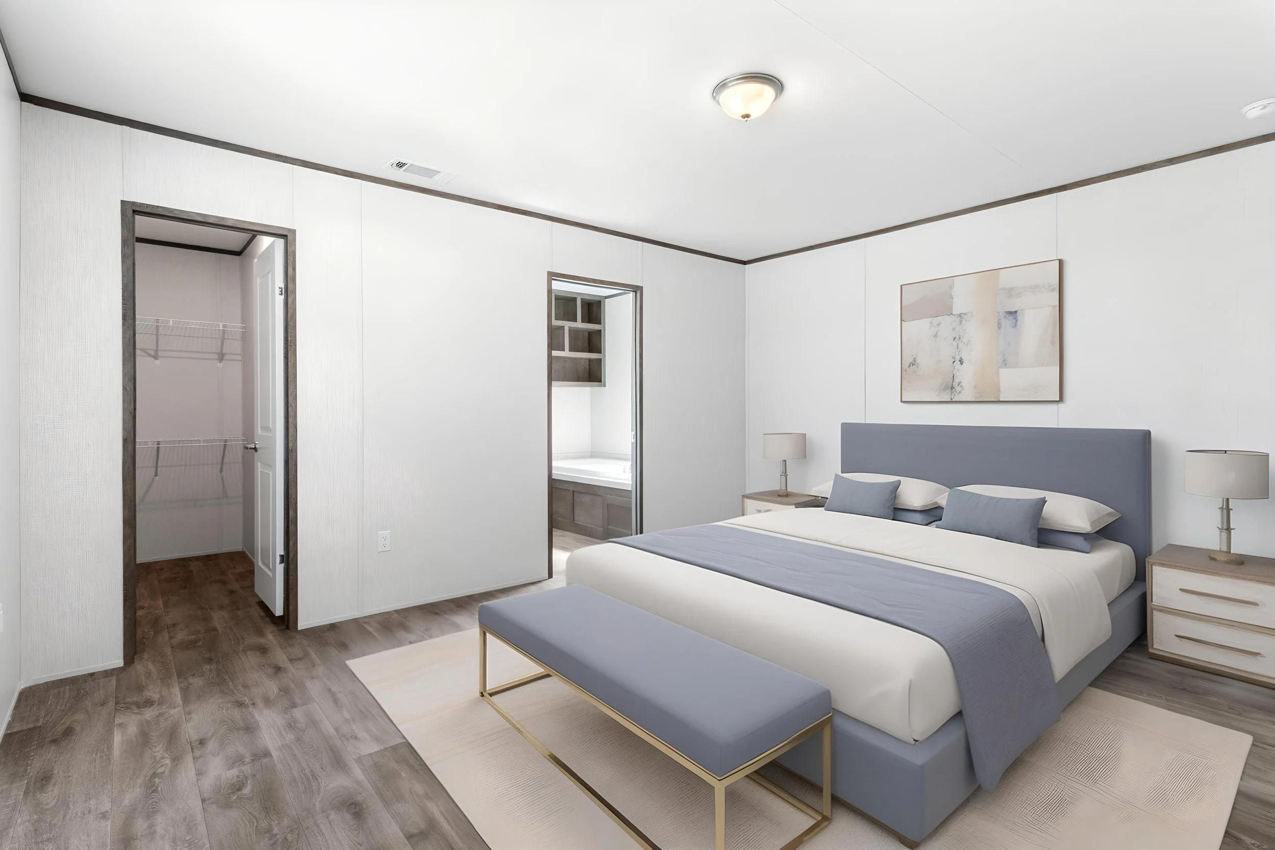 A bedroom with a bed that has a light blue upholstered headboard and matching pillows, two bedside tables with lamps, a piece of abstract art on the wall, a small bench at the foot of the bed, a closet and a bathroom visible in the background.