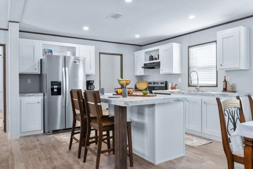 Modern kitchen with white cabinets, stainless steel refrigerator, marble countertop island, wooden chairs, and a window with blinds.