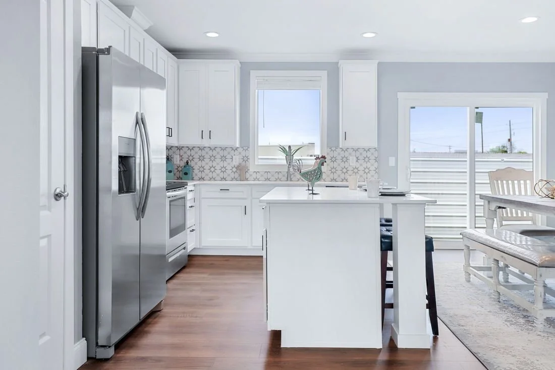 Bright, modern kitchen with white cabinets, stainless steel refrigerator, stove, and patterned backsplash, adjacent to dining area with white table, bench, and sliding glass door.