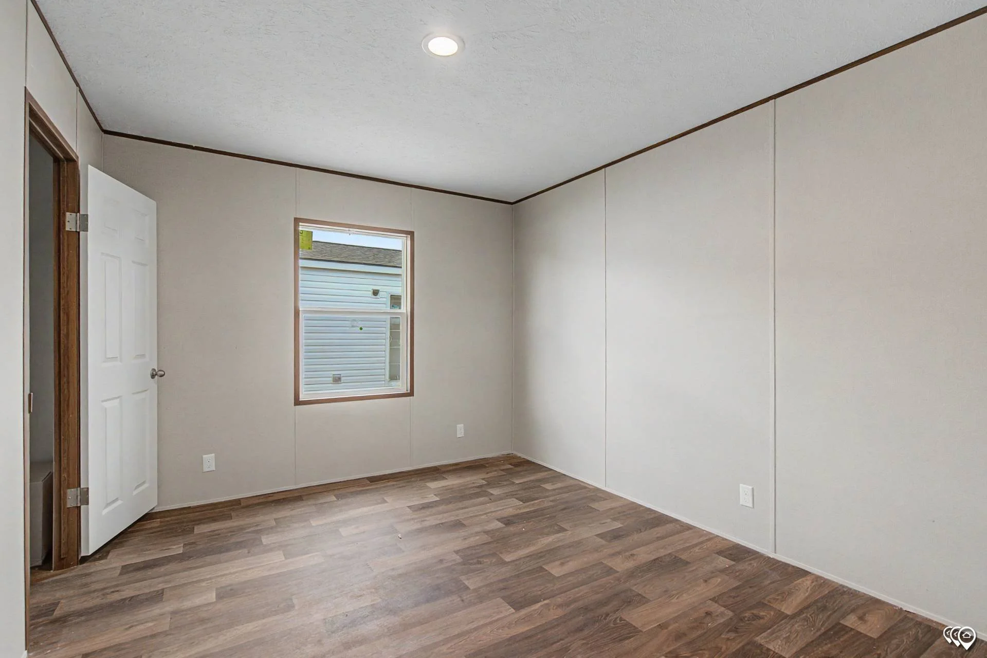 Empty room with light-colored walls, wooden flooring, a door on the left, and a window showing exterior siding and a neighboring house.