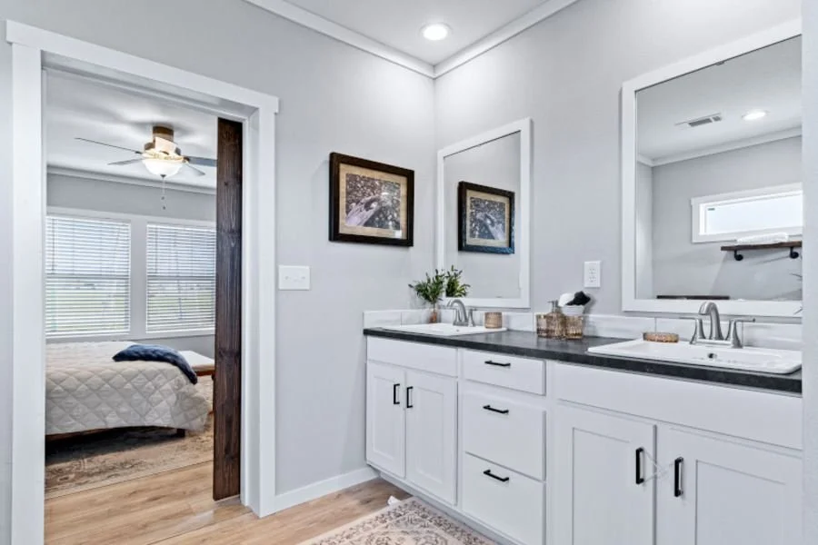 Bathroom with double sink vanity, large mirror, framed pictures, and a view of a bedroom with a bed and windows.