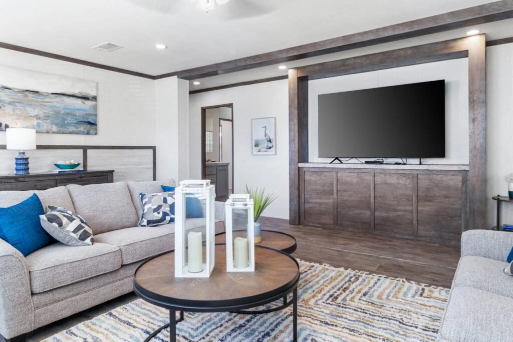Living room with beige sofas, blue and patterned pillows, round wooden coffee table with white lanterns and candles, mounted flat-screen TV, and decorative blue and white accents.