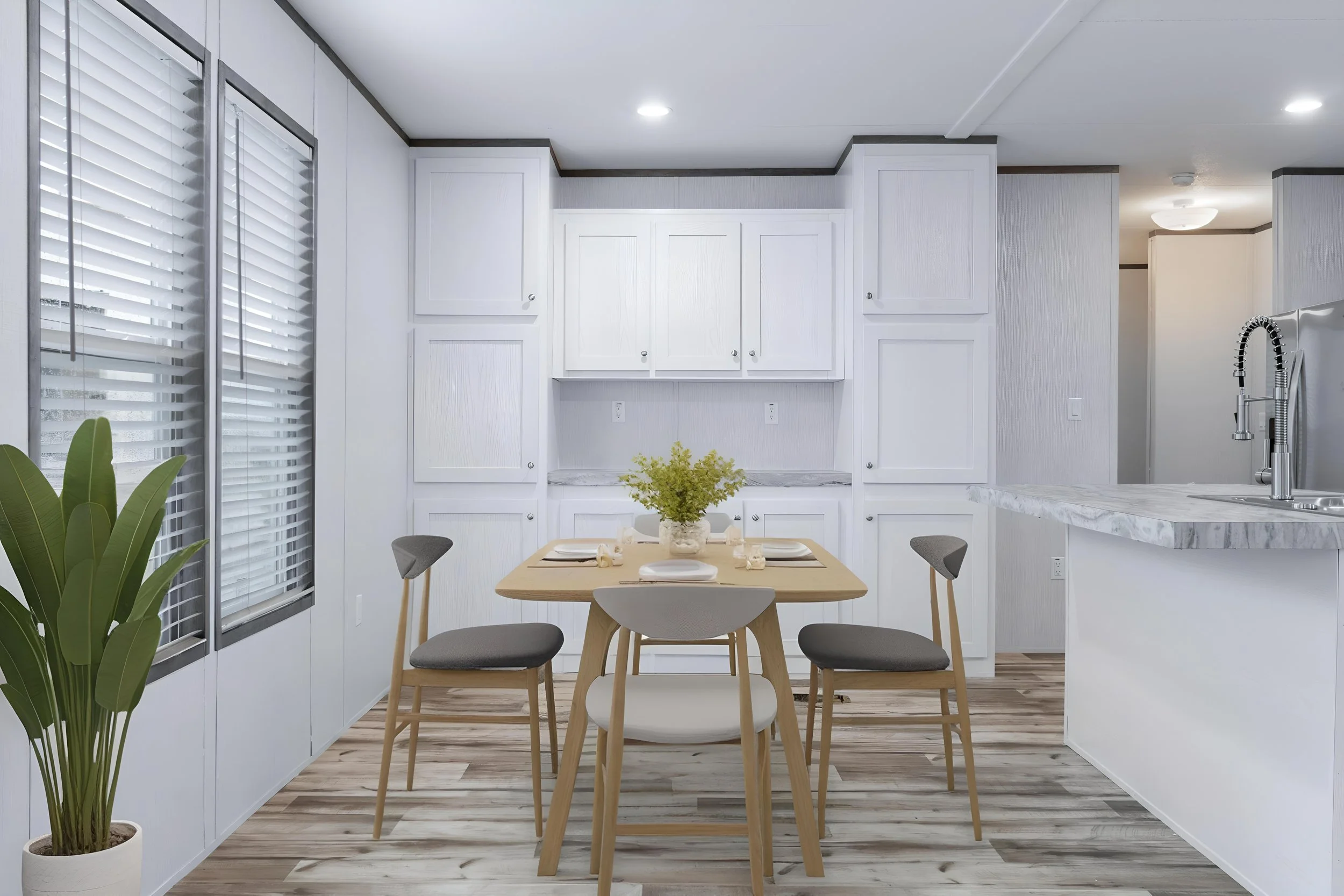 A modern kitchen with white cabinets, a marble countertop, and a dining table with chairs. There are large windows with blinds on the left and a potted plant in the foreground.
