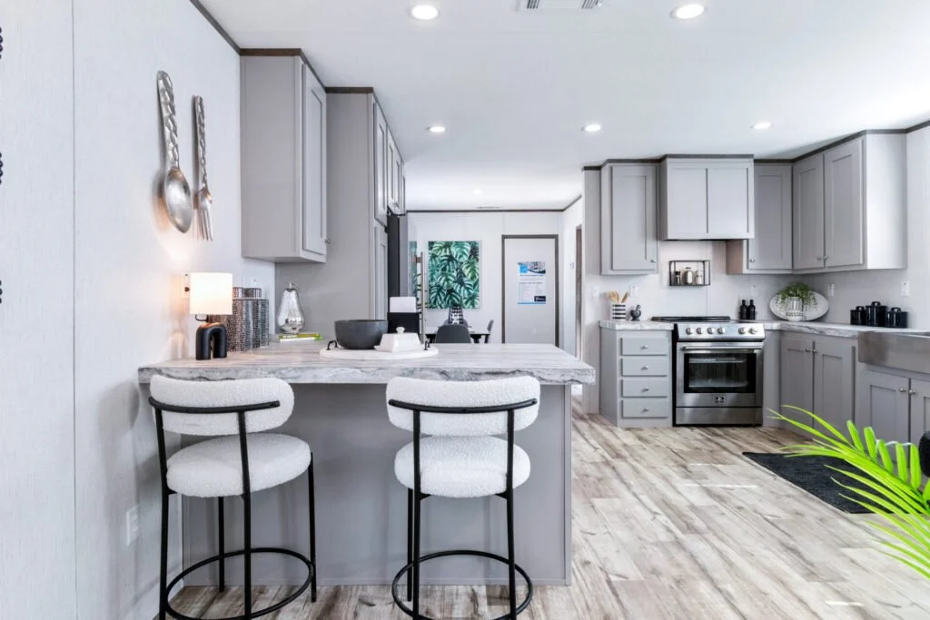 Modern kitchen with light gray cabinets, stainless steel stove, white marble countertop island with two white bar stools, wooden flooring, and decorative items on the counters and wall.