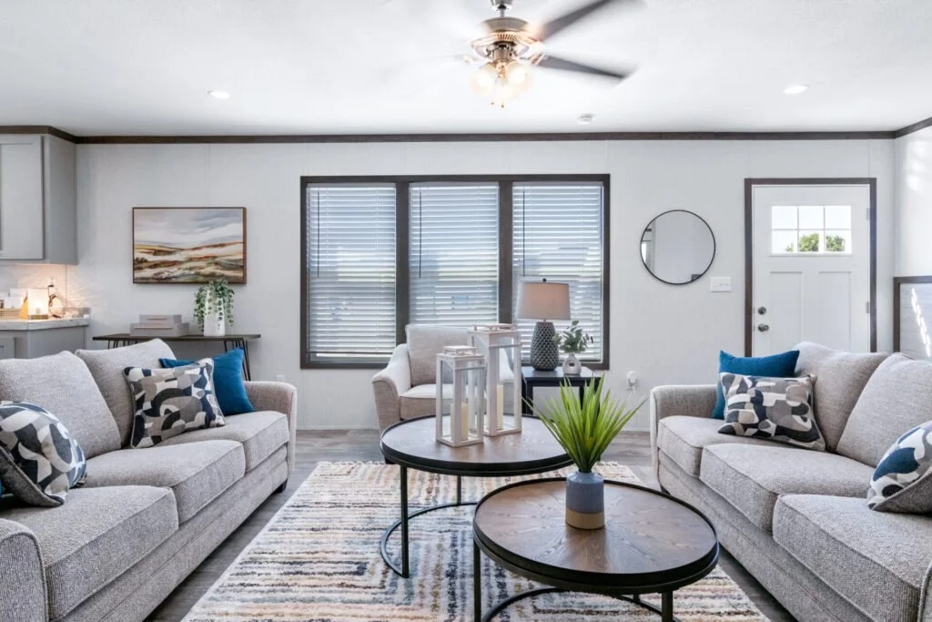 Bright living room with two gray sofas adorned with patterned and blue pillows, centered by two round coffee tables with a plant and decorative lanterns, large windows with blinds, and a white door.