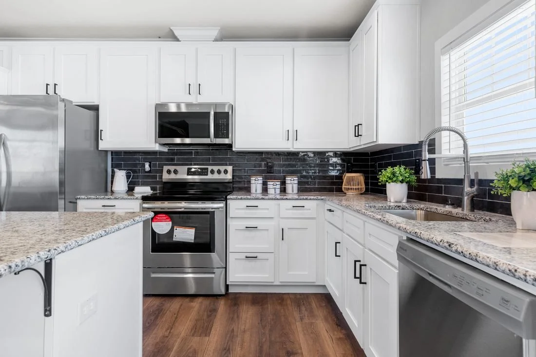 Modern kitchen with white cabinets, granite countertops, stainless steel appliances, black backsplash, and green potted plants.