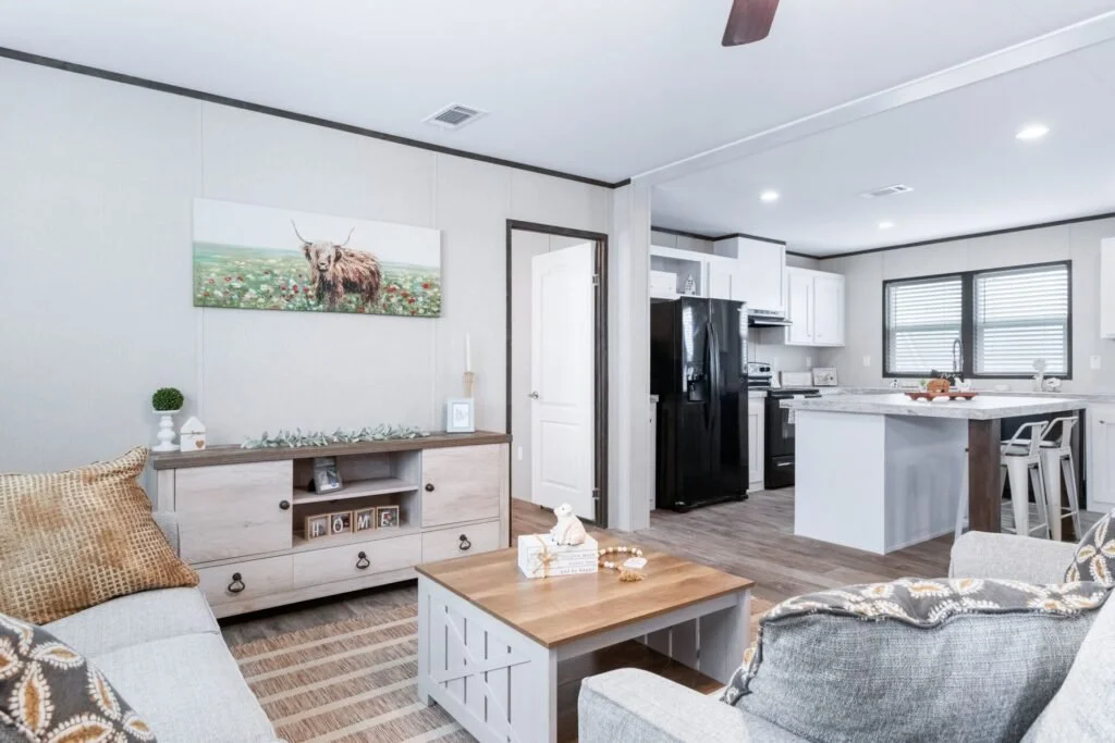 Living room with light-colored sofa, coffee table, and wall-mounted art of a highland cow, connected to a kitchen with white cabinets, black appliances, and a breakfast bar with chairs.