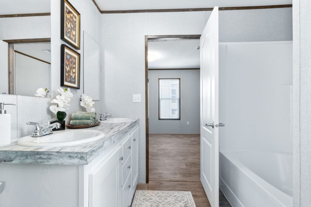 Bathroom with a marble countertop vanity, white sink, soap dispenser, folded green towels, potted white orchids, wall mirror, two framed butterfly art prints, open door revealing a bathtub, and a view of a room with a window and wooden floor.