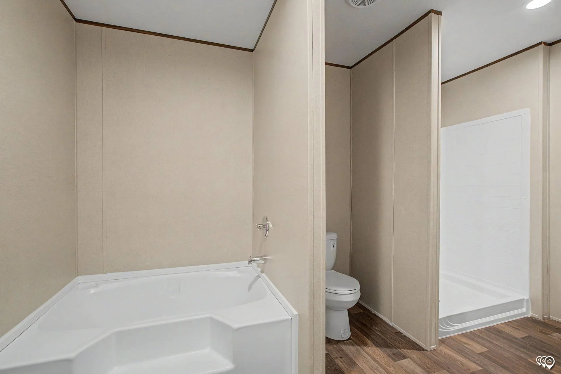 A small bathroom with beige walls, a corner shower with a white tub, a toilet, and a partial wall separating the shower and toilet. The floor has wood-like flooring, and the ceiling has a vent.