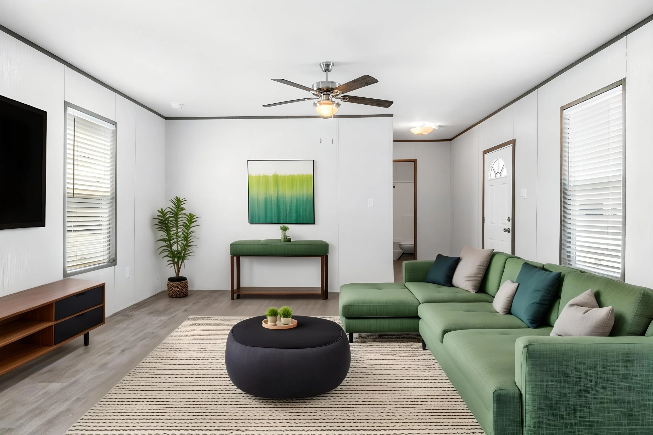 Living room with green sofa, black round ottoman, striped rug, wall art, potted plants, windows with blinds, TV, and ceiling fan.