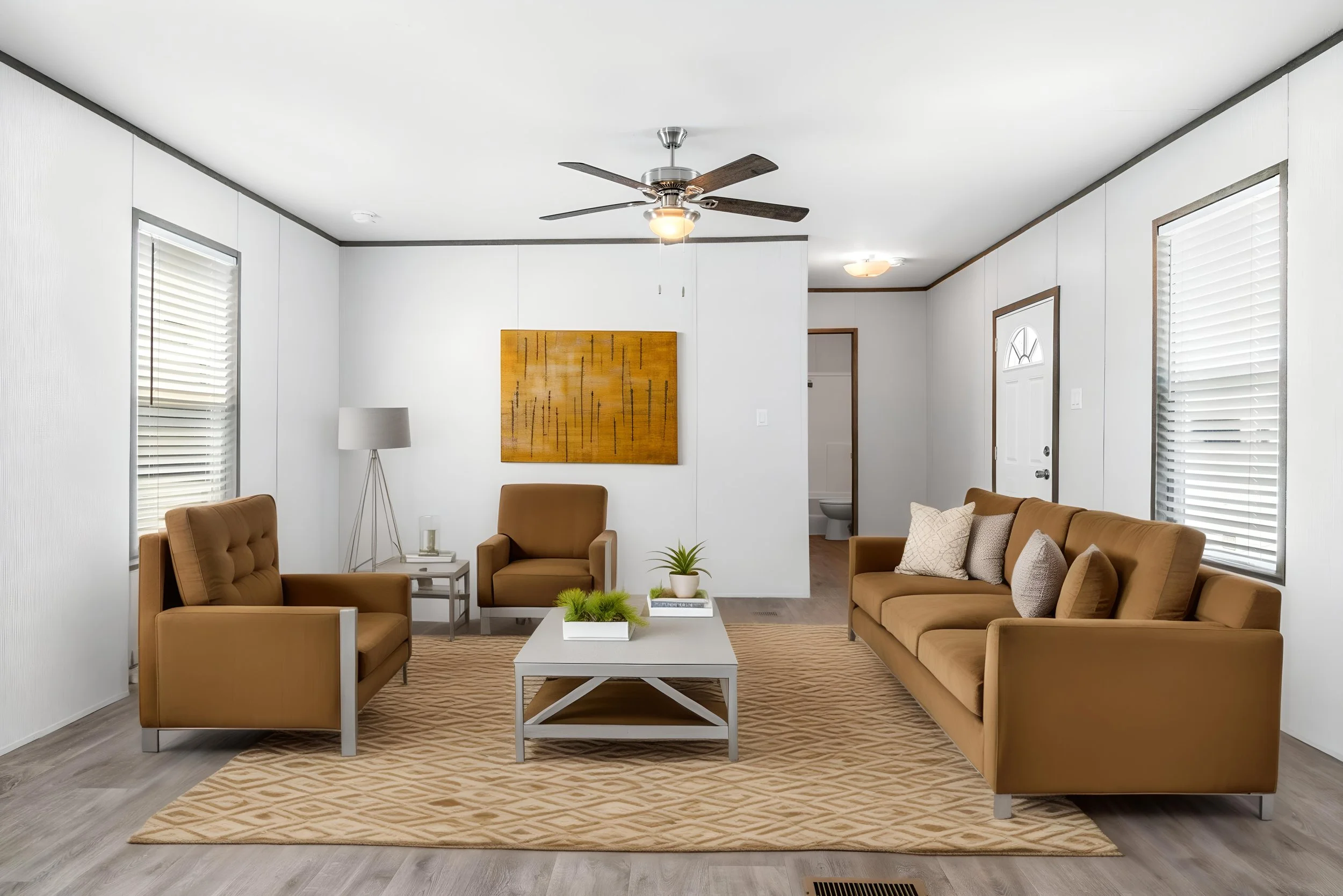 Living room with beige sofas, a white coffee table, a striped area rug, three windows with blinds, a wall art piece, a tall lamp, a ceiling fan, and an open doorway leading to a bathroom.