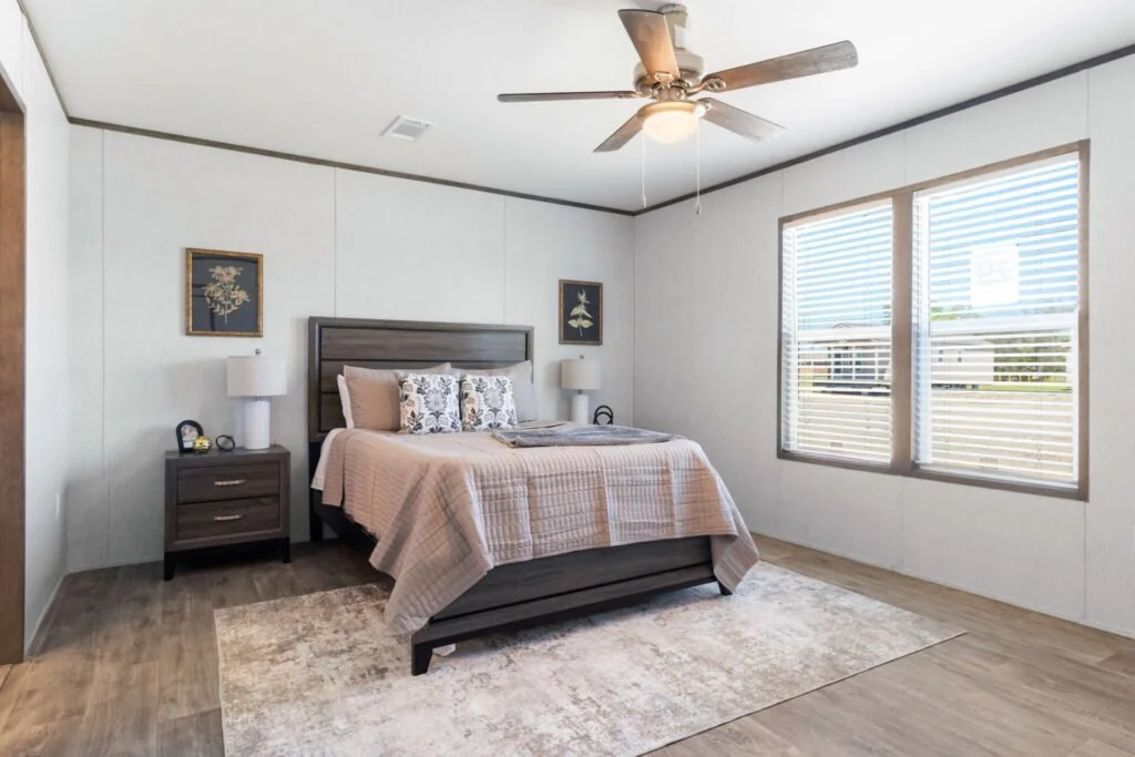 A bedroom featuring a bed with beige and floral pillows, dark wood nightstands with lamps, and a large window with blinds, wood flooring, and a ceiling fan.