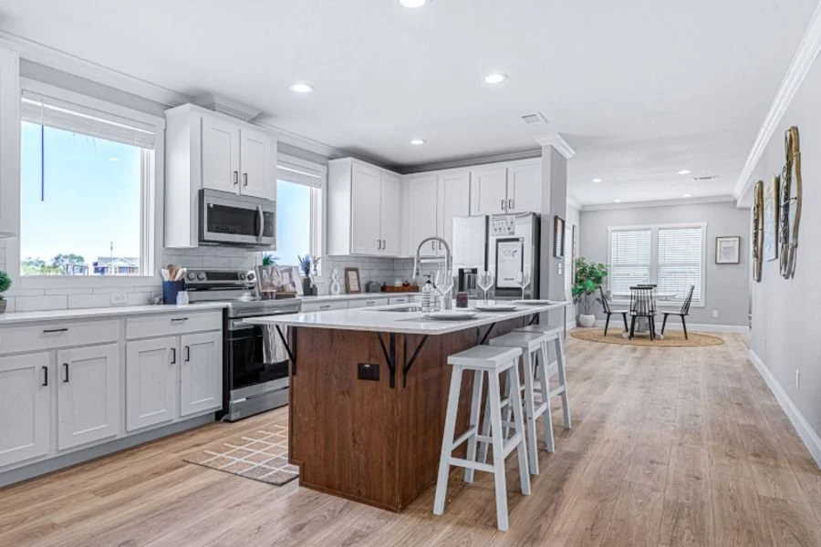 Bright, modern kitchen with white cabinets, stainless steel appliances, a central wooden island with white stools, and a dining area with a round table and black chairs in the background.