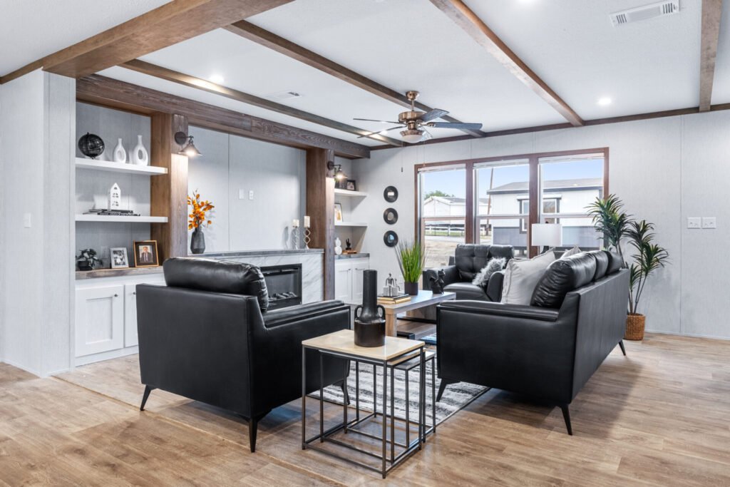 Living room with black leather sofa and armchair, wooden ceiling beams, large window, and decorative items on white shelves.