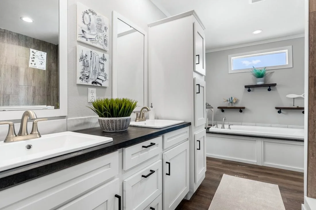 Bathroom with double vanity sinks, white cabinets, black countertop, and a decorative plant in a black and white pot. There is a large mirror above the sinks, artwork on the wall, a bathtub with three floating shelves decorated with plants and orname