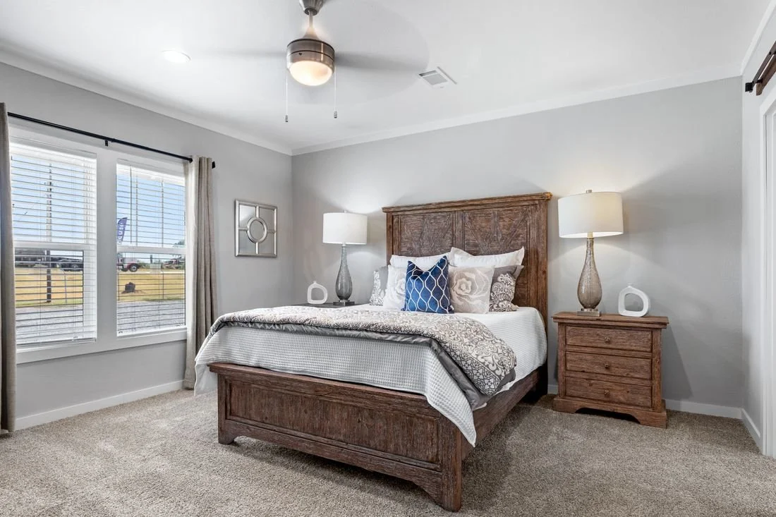 Bedroom with wooden bed, white bedding, multiple pillows, two matching nightstands with lamps, and three windows with blinds and curtains.
