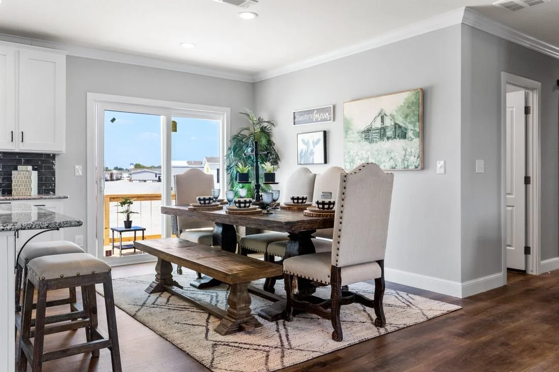 Dining room with a wooden table, six beige chairs, a bench, and a sliding glass door leading to a balcony with a view of neighboring houses and blue sky.