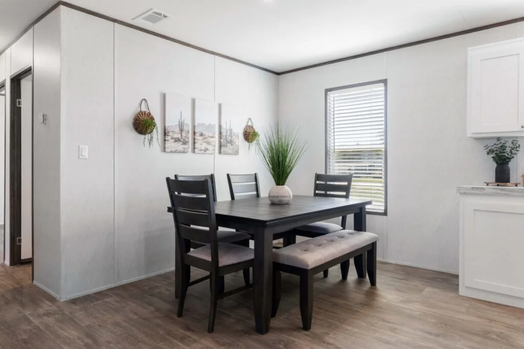 Dining room with a black table, four chairs with beige cushions, a potted green plant, and wall art with hanging baskets. There's a window with blinds and a white kitchen cabinet with a potted plant.