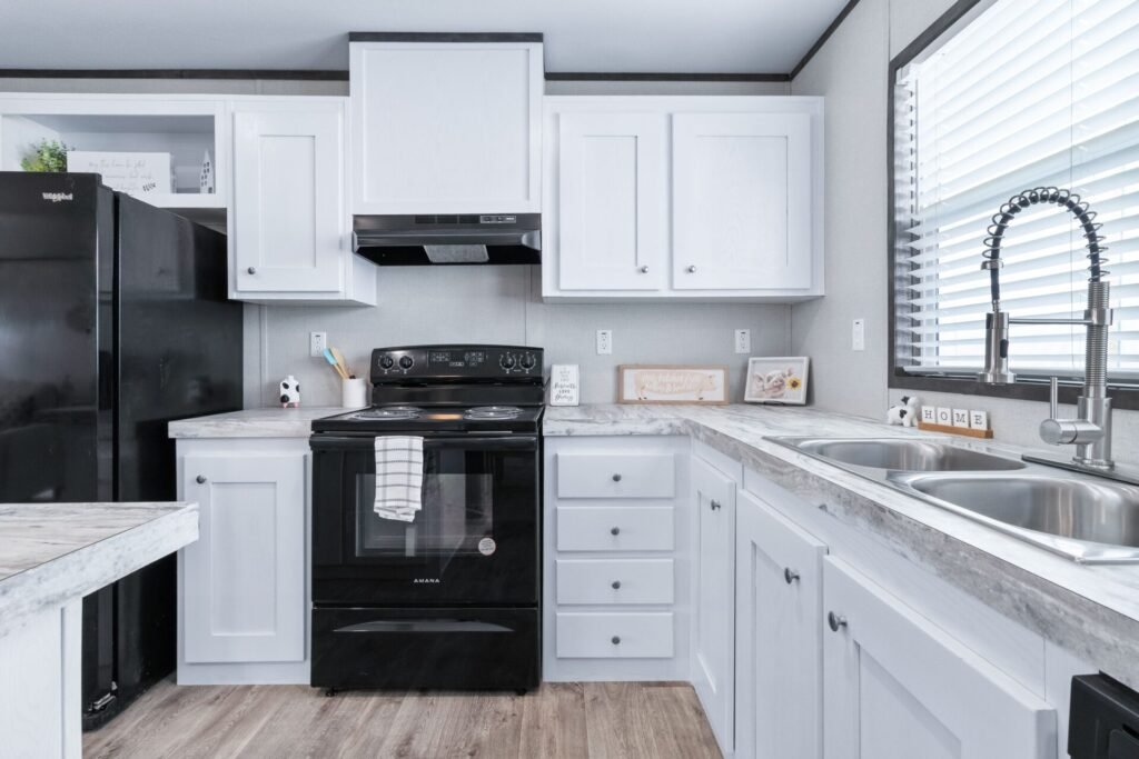 A modern kitchen with white cabinets, a black stove, a silver sink with a faucet, and a black refrigerator. There are small decorative items on the countertop, including a small cow figurine and framed pictures. A window with blinds is above the sink