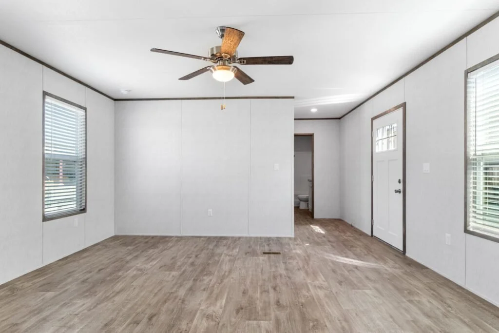 Empty living room with wood flooring, white walls, window blinds, a ceiling fan, and a door leading to a bathroom.