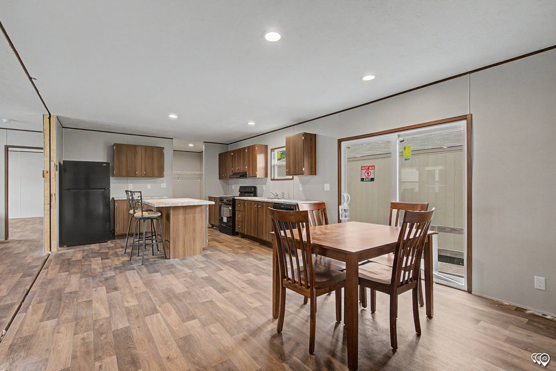 Open-concept kitchen and dining area with wooden cabinetry, a black refrigerator, a kitchen island with stools, and a wooden dining table with chairs. There are sliding glass doors and windows letting in natural light.