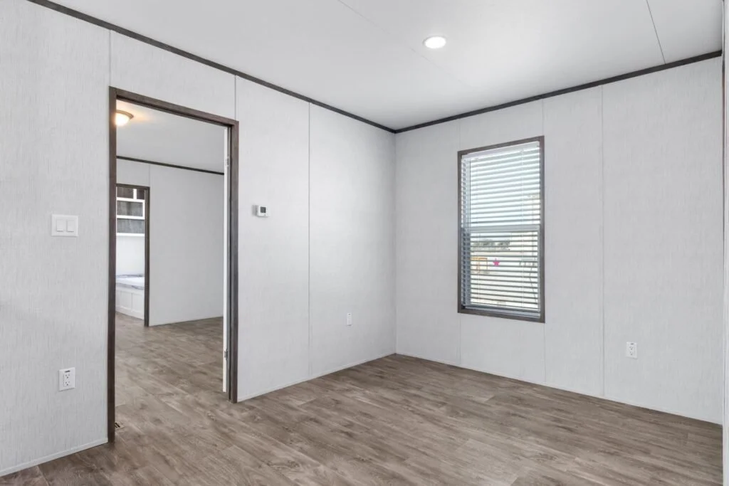 Empty room with white walls, wood laminate flooring, a window with blinds, and a doorway leading to another room.