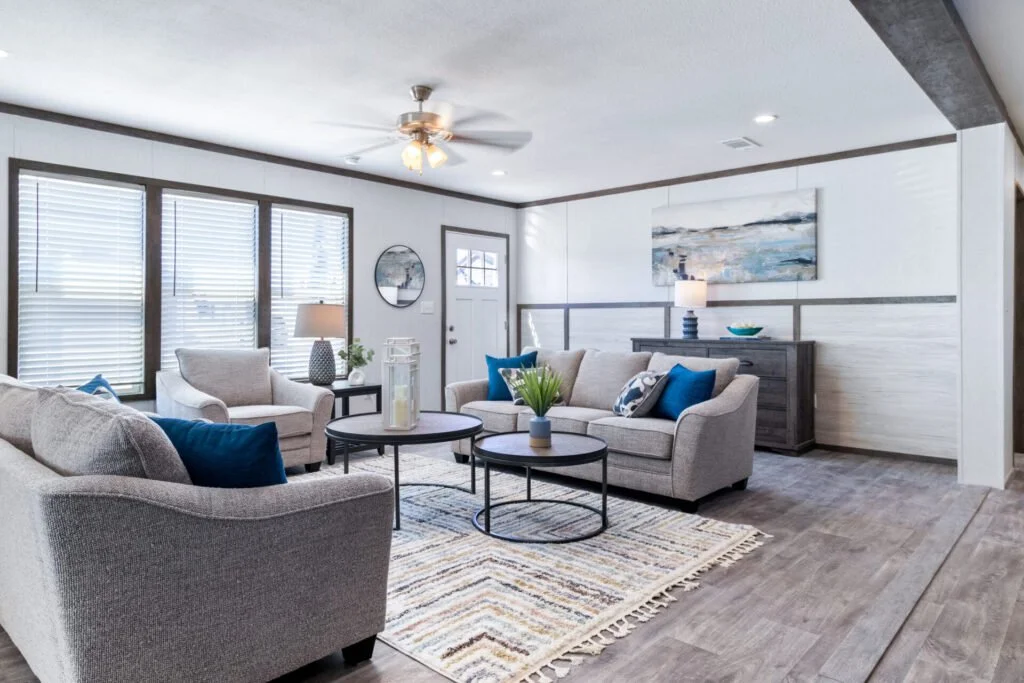 Living room with three beige sofas, blue pillows, a patterned rug, and a coffee table with decorative items, overlooking a wall with windows and a door, in a bright, modern space.